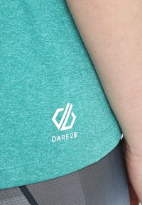 Tyrkysové sportovní tričko s jemnou strukturou a bílým logem "Dare2B" blízko lemu, spárované s černými legínami.