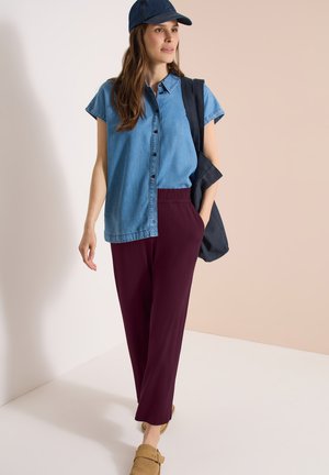 Femme portant une chemise en denim bleu manches courtes, une casquette bleu marine, un pantalon bordeaux, des chaussures beige et portant un sac bleu marine sur l'épaule.