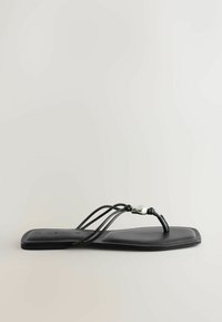 Sandale plate noire avec trois fines brides, une boucle pour l'orteil, et un petit accent métallique sur une des brides, présentée de profil sur un fond neutre.