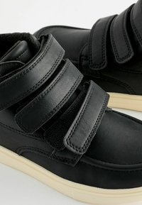 Chaussures en simili cuir noir avec trois bandes Velcro, bout rond et semelle en caoutchouc clair. Surface texturée et détails de couture visibles.