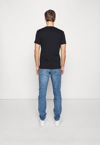 Svart t-shirt med kort ärm, kombinerad med ljusblåjeans och vita sneakers, visad bakifrån mot en enfärgad bakgrund.