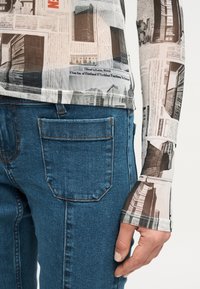 Κοντινό πλάνο ενός ατόμου που φοράει μπλε τζιν και μακρυμάνικη μπλούζα με ασπρόμαυρη εκτύπωση εφημερίδας.