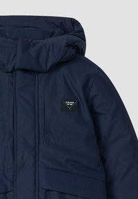 Donkerblauwe gewatteerde jas met een hoge kraag, voorzien van een logo patch op de borst, horizontale naaddetails en een gladde textuur.