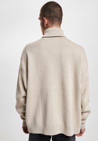 Homme portant un pull en maille beige à col roulé, de dos, avec des cheveux courts et foncés et un pantalon noir, sur fond blanc uni.