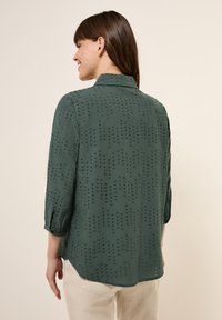 Blouse vert foncé en tissu léger, avec des détails en œillets et des manches trois-quarts, le dos a un ourlet arrondi et un col.