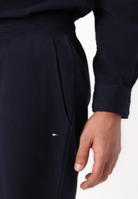 Højre hånd ved siden af lommen på sorte joggingbukser med lille rød, hvid og blå rektangulært logo på låret, iført en matchende sort langærmet top.