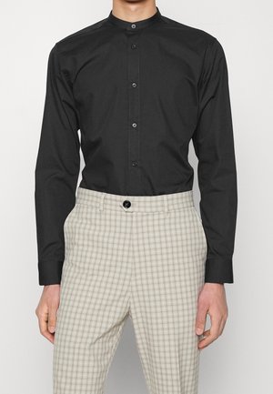 Homme portant une chemise noire à col mandarin boutonnée rentrée dans un pantalon beige clair à carreaux avec un bouton noir.