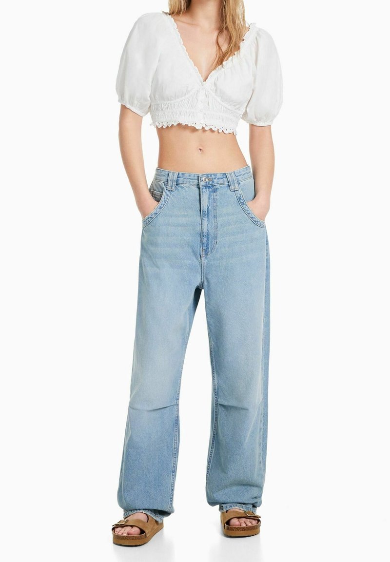 Femme portant un crop top blanc à manches bouffantes, un jean bleu clair à jambes larges, les mains dans les poches, et des sandales plateformes marron sur fond blanc.