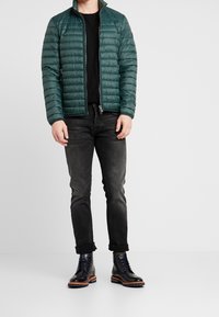 Mørkegrøn pufferjakke, quilted design, lynlåslukning, båret over en sort skjorte, parret med sorte jeans og navy støvler med orange detaljer.
