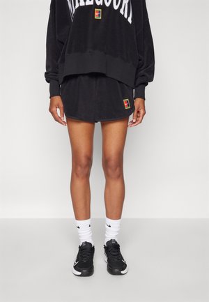 Personne portant un sweat-shirt noir oversize et un short assorti avec des logos colorés, des chaussettes blanches et des chaussures de sport noires, debout.