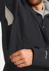 Schwarze Jacke mit einer Reißverschlusstasche auf der Brust, ausgestattet mit einem weichen Innenfutter und verstellbaren Manschetten. Das Material scheint leicht und langlebig zu sein.