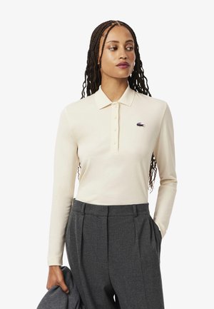 Cremfarbener Langarm-Polo mit Kragen und fünf Knöpfen, ausgestattet mit einem kleinen lila Lacoste-Logo auf der linken Brust, kombiniert mit grauen Hosen.