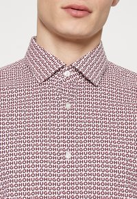 Lyhythihainen valkoinen paita, jossa toistuva punainen ja musta "HUGO"-kuvio, klassinen kaulus ja napitus. Sileä kangaspinta.
