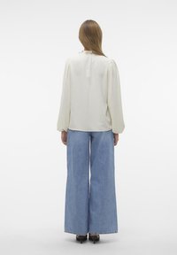 Blouse blanche texturée avec des manches bouffantes, associée à un jean bleu à jambes larges et des talons noirs. La vue arrière montre un subtil détail volanté au niveau de l'encolure.