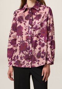 Long-sleeve blouse in pink fabric with large purple floral pattern, button closure, and pointed collar. Smooth texture, relaxed fit.