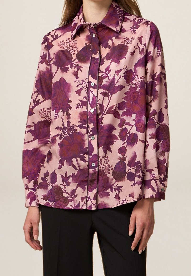 Long-sleeve blouse in pink fabric with large purple floral pattern, button closure, and pointed collar. Smooth texture, relaxed fit.