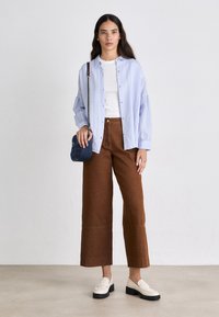 Lichtblauwe gestreepte overhemd met knopen over een wit T-shirt, gecombineerd met bruine wijde broek en witte loafers; houdt een blauw gestreepte handtas.