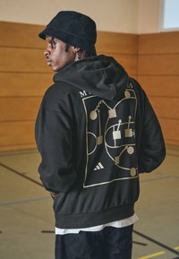 Hanorac negru cu glugă, cu un imprimeu grafic aurit pe spate, prezentând designuri abstracte și logo-ul Adidas. O strat vizibil de cămașă albă dedesubt.
