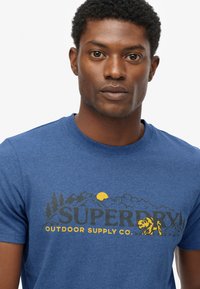 Mand med krøller iført en blå Superdry T-shirt med gult og sort bjerg, sol, træer og tigerlogo på hvid baggrund.