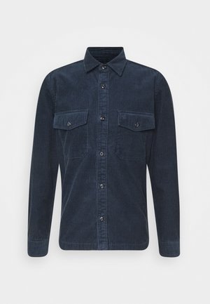 Camicia a maniche lunghe in velluto a coste blu navy con chiusura frontale con bottoni e tasche sul petto con patta e bottone, mostrata su uno sfondo semplice.
