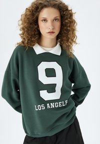 Ciemnozielona bluza z białym kołnierzykiem, z dużą białą cyfrą "9" w czarnym obramowaniu i napisem "LOS ANGELES" w białym kolorze poniżej.