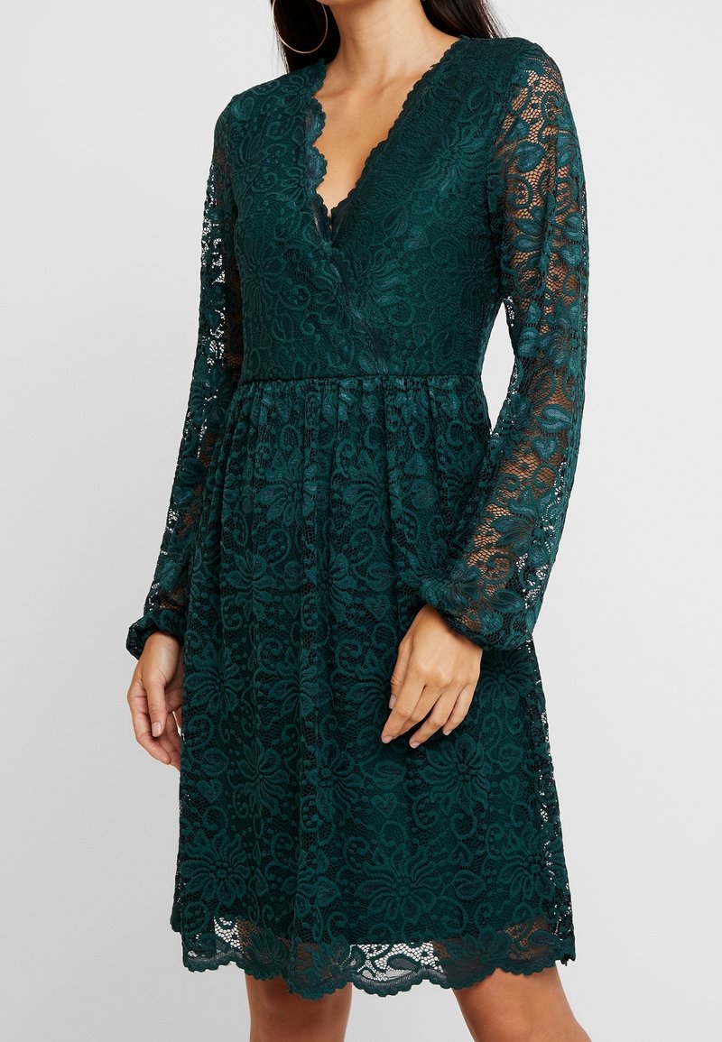 Femme portant une robe en dentelle vert foncé avec de longues manches transparentes et un décolleté en V festonné, debout devant un fond uni.