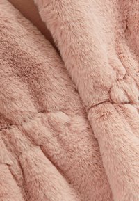 Matériau en fausse fourrure rose clair avec une texture douce et duveteuse. Le design présente des sections rembourrées et en relief, ajoutant un intérêt visuel.