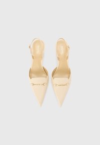 Paire de ballerines beige à bout pointu avec boucle dorée et brides à la cheville, vues de dessus sur fond gris clair.