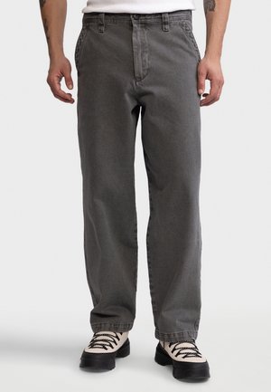 Homme portant un pantalon ample gris foncé et des bottes à lacets noires et blanches, debout devant un fond uni.