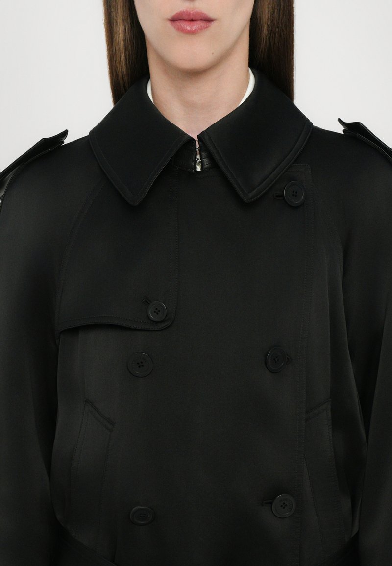 Cappotto nero lungo con colletto ampio, frontale doppiopetto, bottoni e una texture elegante. Presenta una tasca laterale e dettagli con zip.