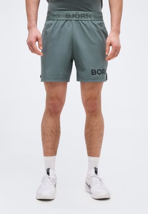 Bărbat purtând pantaloni scurți atletici gri Björn Borg, șosete albe până la gleznă și adidași albi Asics, stând cu picioarele ușor depărtate pe un fundal alb.