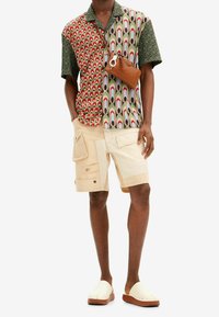 Kortärmad skjorta med flerfärgade geometriska mönster; beige cargoshorts med fickor; vita slip-on-skor; brun axelremsväska.