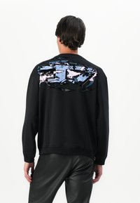 Homme portant un sweat-shirt noir avec un grand motif abstrait bleu et rose dans le haut du dos, associé à un pantalon en cuir noir, vu de dos.