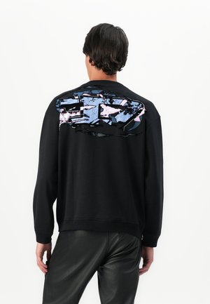 Homme portant un sweat-shirt noir avec un grand motif abstrait bleu et rose dans le haut du dos, associé à un pantalon en cuir noir, vu de dos.