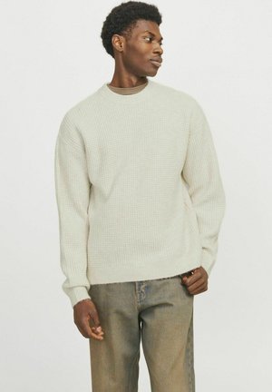 Light beige knitted sweater with a round neckline, long sleeves, and a textured pattern. Paired with loose-fitting, faded denim pants.