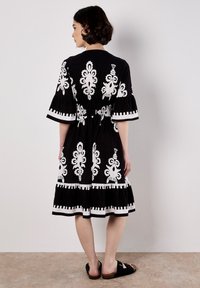 Une femme se tient debout, de dos, dans une robe noire avec des motifs brodés blancs et un ourlet en couches. Elle porte des sandales noires sur un fond neutre.