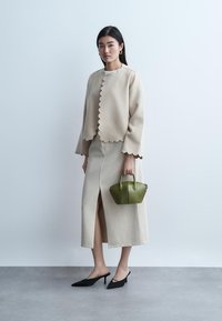 Sieviete tērpusies bēša krāsas vilcienu apmales jaku un svārkos ar priekšējo šķēlumu, turot nelielu zaļu somiņu, un valkā melnus arku papēžus.