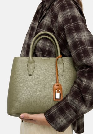 Kobieta trzymająca teksturowaną oliwkowozieloną torebkę z beżową skórzaną metką i srebrną kłódką, ubrana w ciemną kratkowaną kurtkę.