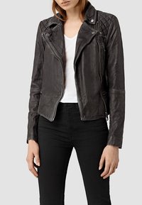 Giacca da motociclista in pelle nera con accenti trapuntati sulle spalle, chiusura con zip asimmetrica e design aderente. Indossata sopra una t-shirt bianca.