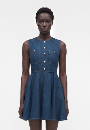 Γυναίκα με κοντά μαλλιά, ντυμένη με μπλε φόρεμα από τζιν χωρίς μανίκια, με χρυσά κουμπιά και τσέπες στο στήθος, που στέκεται μπροστά σε απλό λευκό φόντο.