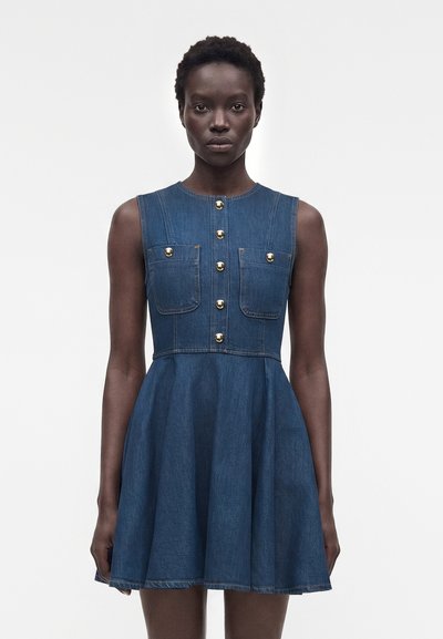 Femme aux cheveux courts portant une robe en denim bleue sans manches avec des boutons dorés et des poches poitrine, debout devant un fond blanc uni.