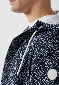 Primo piano di una persona che indossa una giacca blu navy con motivo intrecciato bianco "JS", stringhe bianche e una toppa rotonda con logo sulla manica.