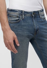 Primo piano di una persona che indossa jeans di denim blu e una camicia bianca, con la mano sinistra rilassata lungo il fianco.