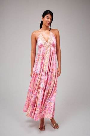 Vrouw staande, gekleed in een roze maxi-jurk met halternek en patroon, en gouden slippers, tegen een effen grijze achtergrond.