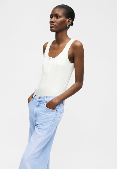 Tume nahaga naine, kes kannab valget varrukateta topi ja helesiniseid kõrge vöökohaga teksaseid, seisab käed taskutes ühevärvilise tausta ees.