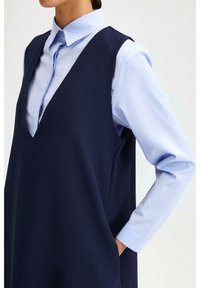 Robe sans manches bleu marine avec un col en V, associée à une chemise col léger bleu clair. Le tissu semble lisse avec un design structuré et une poche visible.
