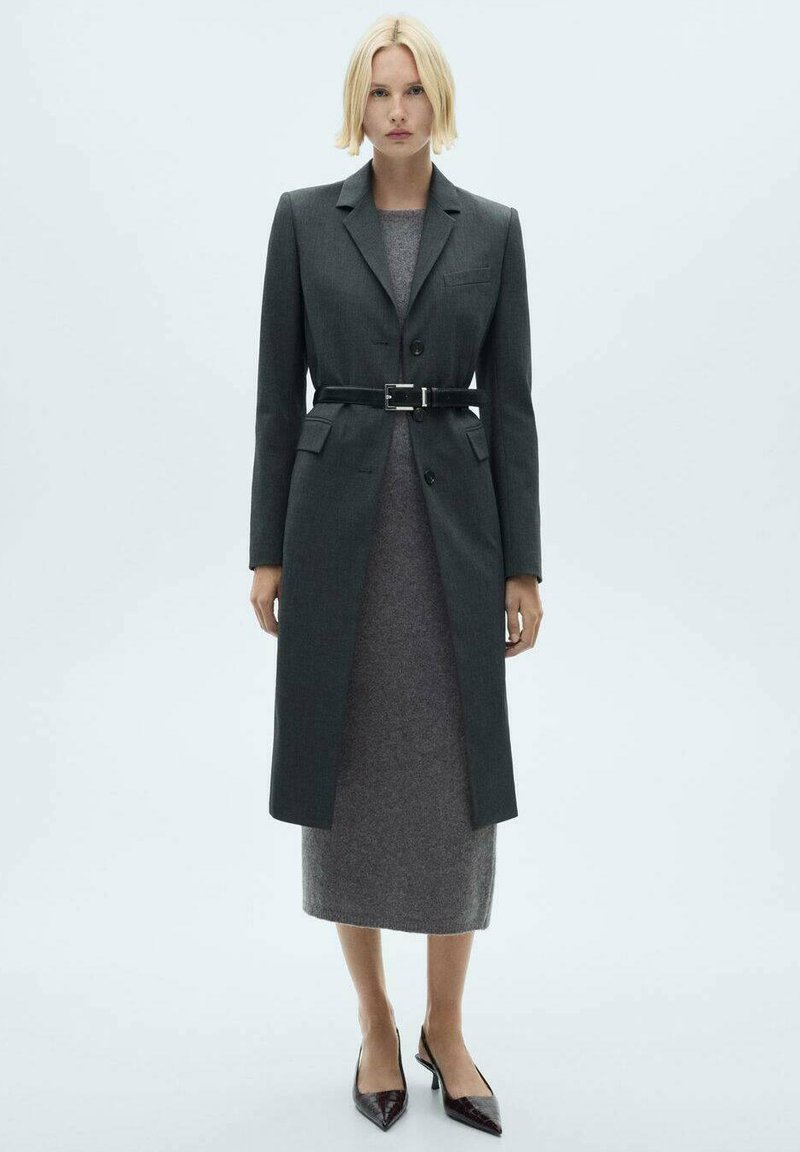 Manteau gris ajusté avec un design structuré, fermeture à un bouton et ceinture noire. Porté par-dessus une robe midi gris foncé, associé à des escarpins à bouts pointus.