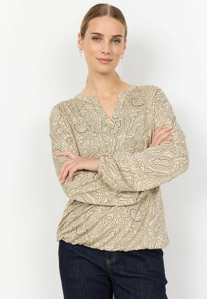 Beige paisley patterned blouse with a V-neckline, long sleeves, and gathered detail at the hem. Paired with dark blue jeans.