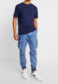 Navy T-shirt with white square patterns, paired with light blue cargo pants featuring side pockets and elastic cuffs, worn with white sneakers.