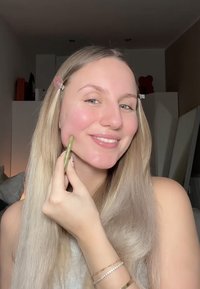 Une femme aux longs cheveux blonds sourit tout en utilisant un outil facial vert sur sa joue. Elle porte un haut léger et a la peau claire.